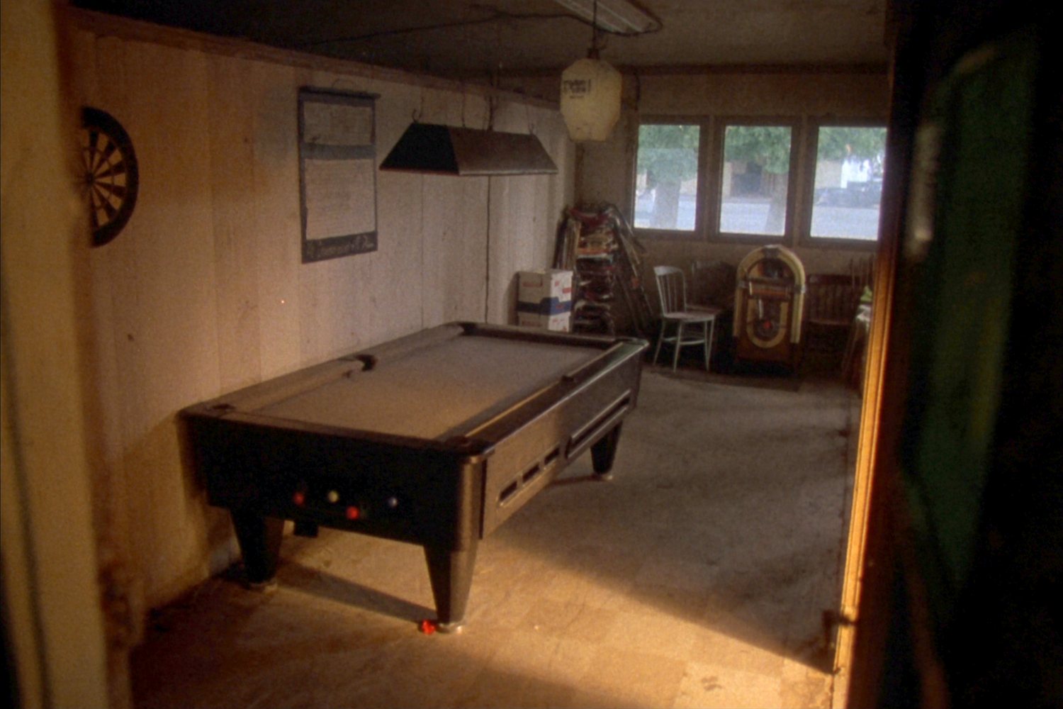 The back game room seen through its doorway: a coin operated pool table under a hanging lamp, a dartboard on the panelled wall, an old jukebox in the corner and daylight through the back windows.