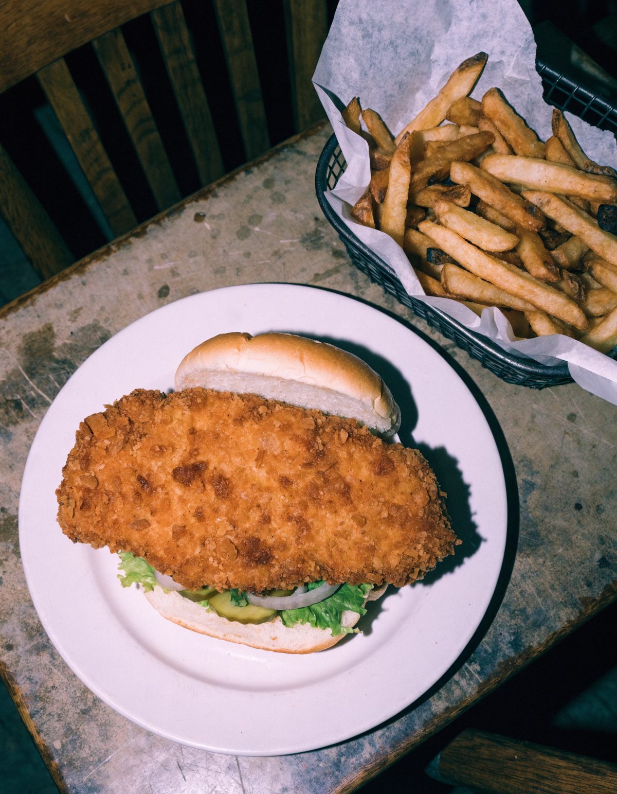 The Mainliner on a plain white plate: a hand breaded pork tenderloin hanging several inches past the bun on every side, with lettuce, pickle and onion under it and a basket of hand cut fries beside it.