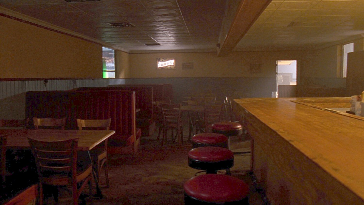 Looking down the long room from just inside the front door: the wooden bar and its red stools along the right, booths and tables along the left, pressed tin ceiling, a lit sign on the back wall and a doorway at the far end.